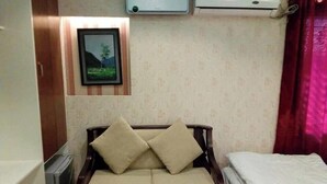 Een kleine kamer met een bed, een bank met kussens, een schilderij aan de muur, en een airconditioning.