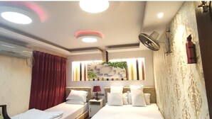 Hotelkamer met twee bedden, een raam met decoratieve wand, een airconditioner, een plafondventilator en een brandblusser.