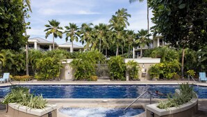A swimming pool surrounded by palm trees and a building with balconies. - New! Stylish island living, walk to beach, opp to Hilton! Partial ocean views! (Waikoloa Village)