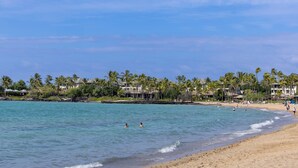 A beach with clear turquoise water, palm trees, and beach houses in the background. - New! Stylish island living, walk to beach, opp to Hilton! Partial ocean views! (Waikoloa Village)