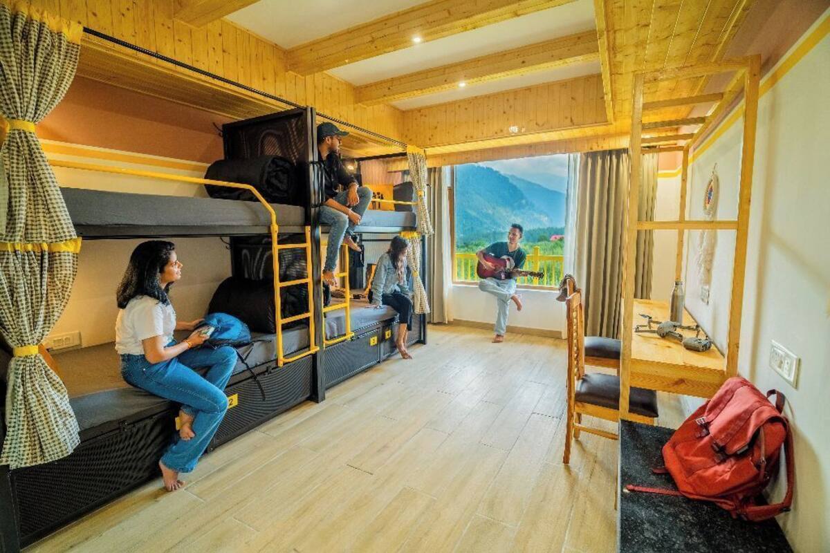 A dormitory room with bunk beds, a wooden interior, a window with a mountain view, and a table with a red backpack.