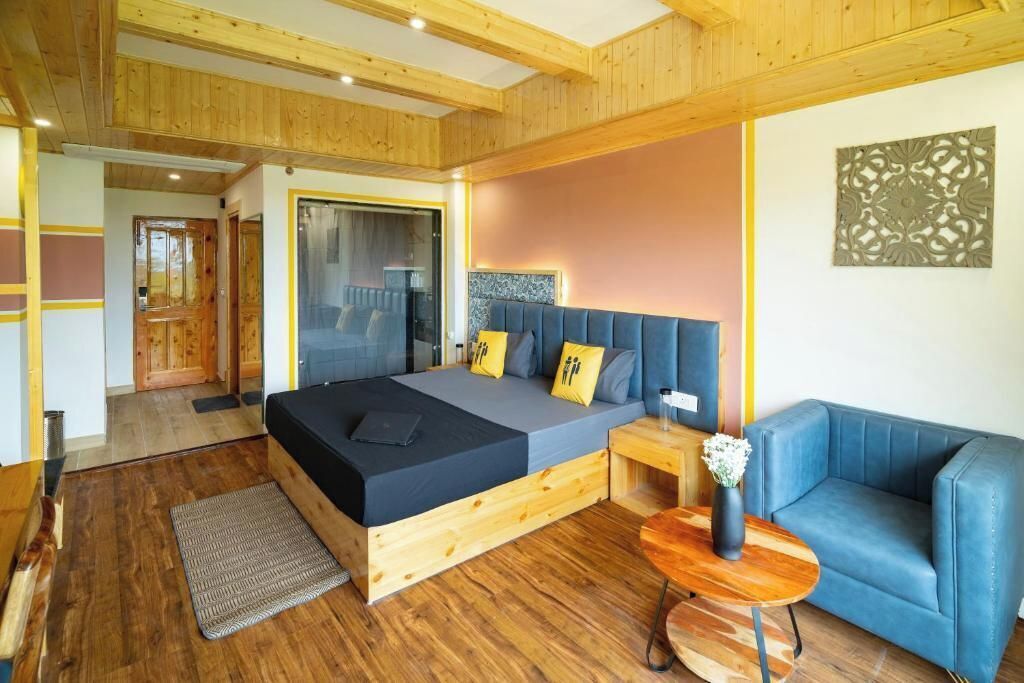 Une chambre moderne avec un plafond en bois, un lit avec un matelas gris, un canapé bleu et une petite table d’appoint en bois sur laquelle est posé un vase.