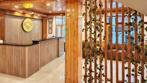 A wooden reception desk with a clock and framed certificates, a wooden partition with climbing plants, and a view of the outside through large windows. - Rosewood Manali (Manali)
