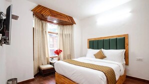 A hotel room with a wooden headboard, a large bed with white bedding, a small bedside table with a vase of flowers, and a window with curtains. - Rosewood Manali (Manali)