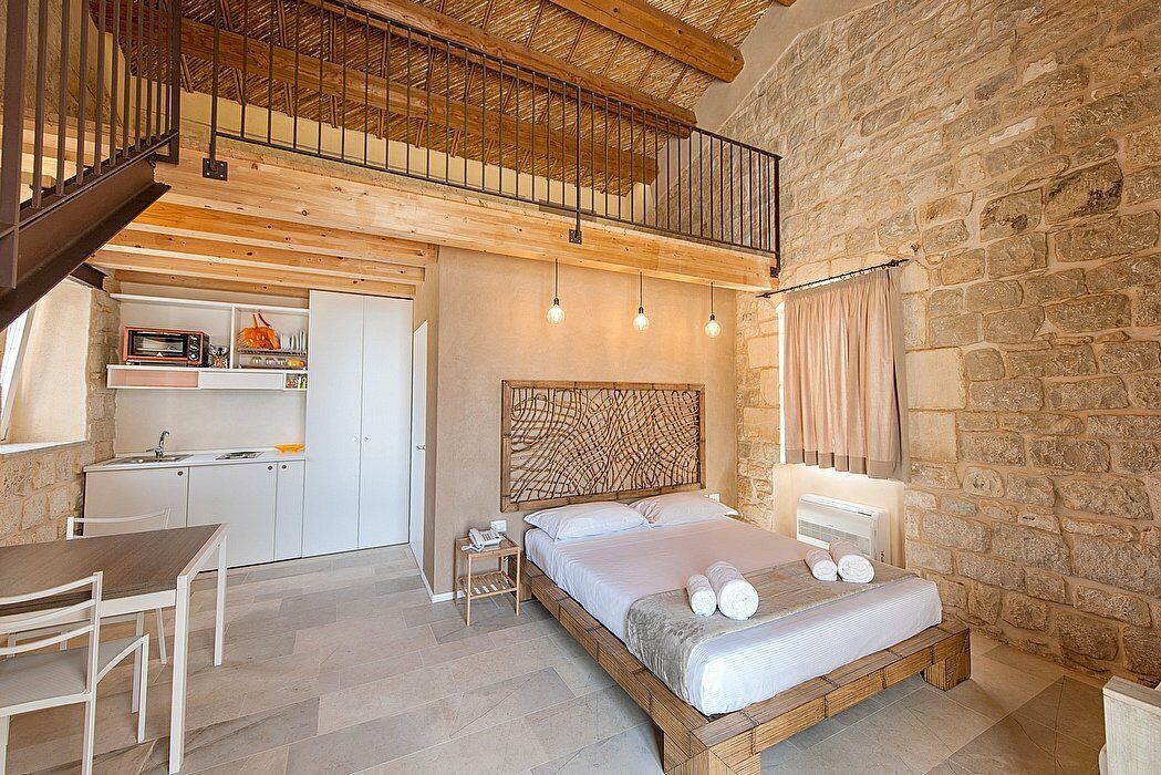 A bedroom with a wooden bed, a kitchen area, and a stone wall.