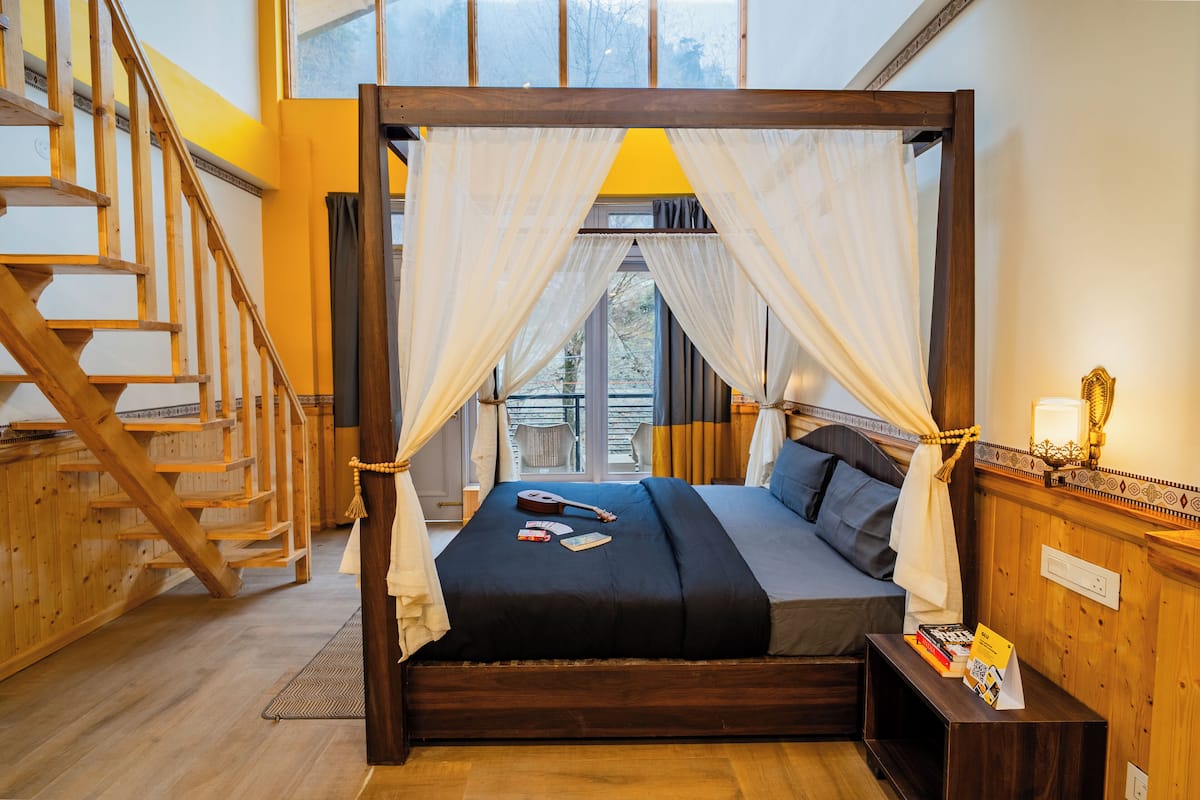 A bedroom with a four-poster bed, wooden flooring, a staircase, and a view through large windows.