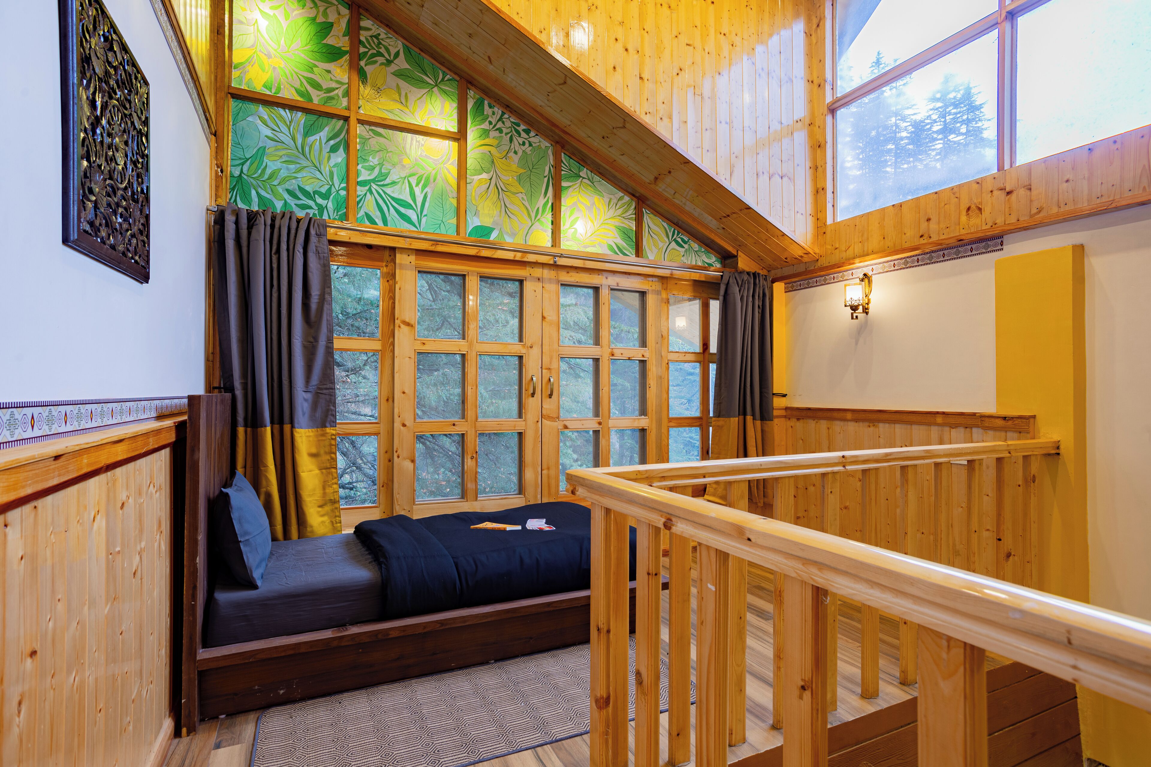 A room with a bed, a window with a view of trees, a wooden railing, and a decorative wall piece.