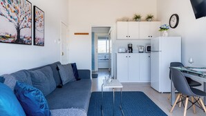 A modern living room with a grey sofa, a small table with glasses, and a kitchen area with white cabinets and appliances. - Lazuli 1-BR W Pool & Balcony in Larnaca (Oroklini)
