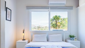 A hotel room with a bed, a nightstand with a lamp, a window with a view of palm trees, and a framed quote on the wall. - Lazuli 1-BR W Pool & Balcony in Larnaca (Oroklini)