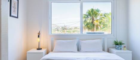 Une chambre d’hôtel avec un lit, une table de chevet avec une lampe, une fenêtre donnant sur des palmiers et un cadre avec une citation accroché au mur.