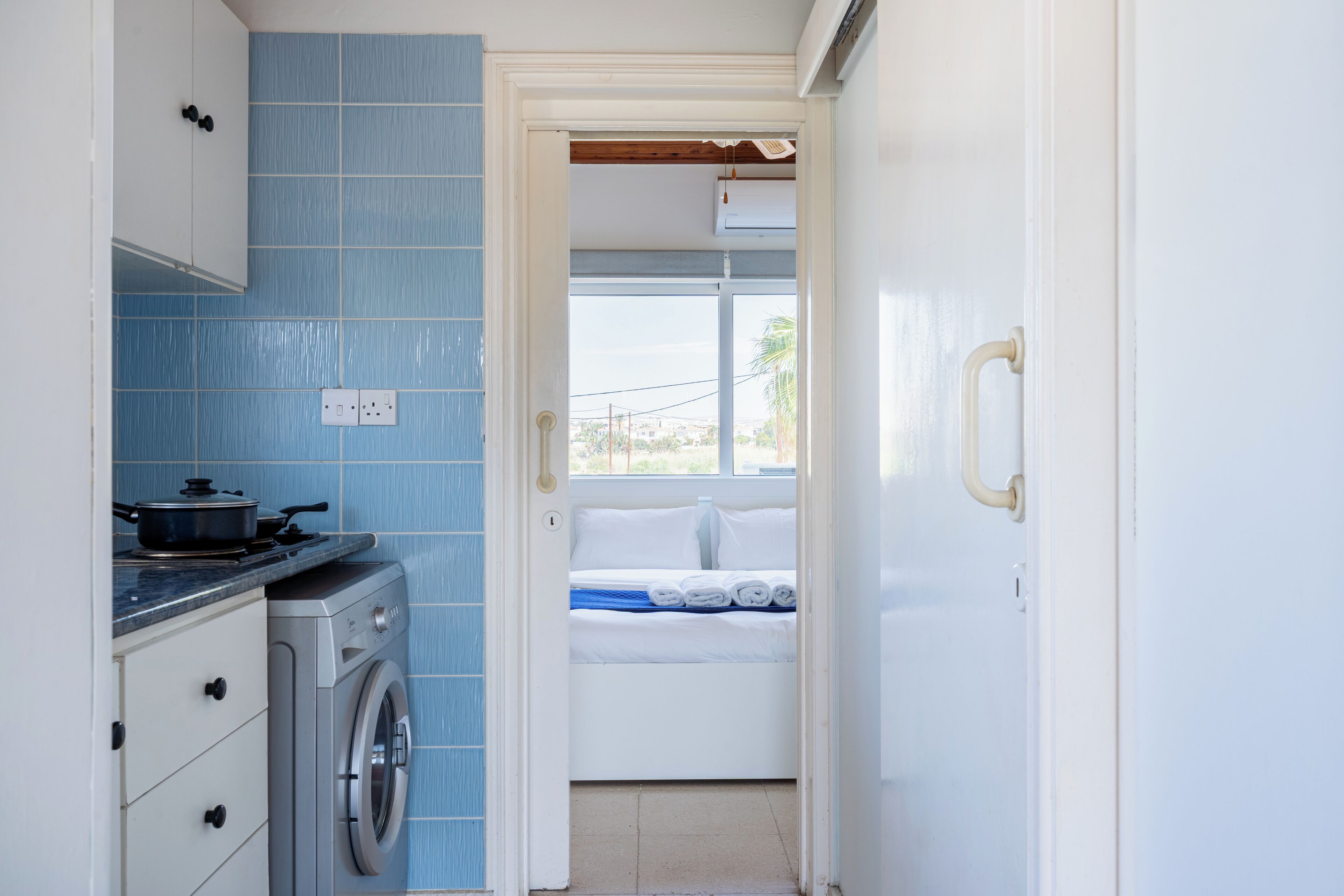A compact kitchen with a stove, washing machine, and a bed visible through an open door.
