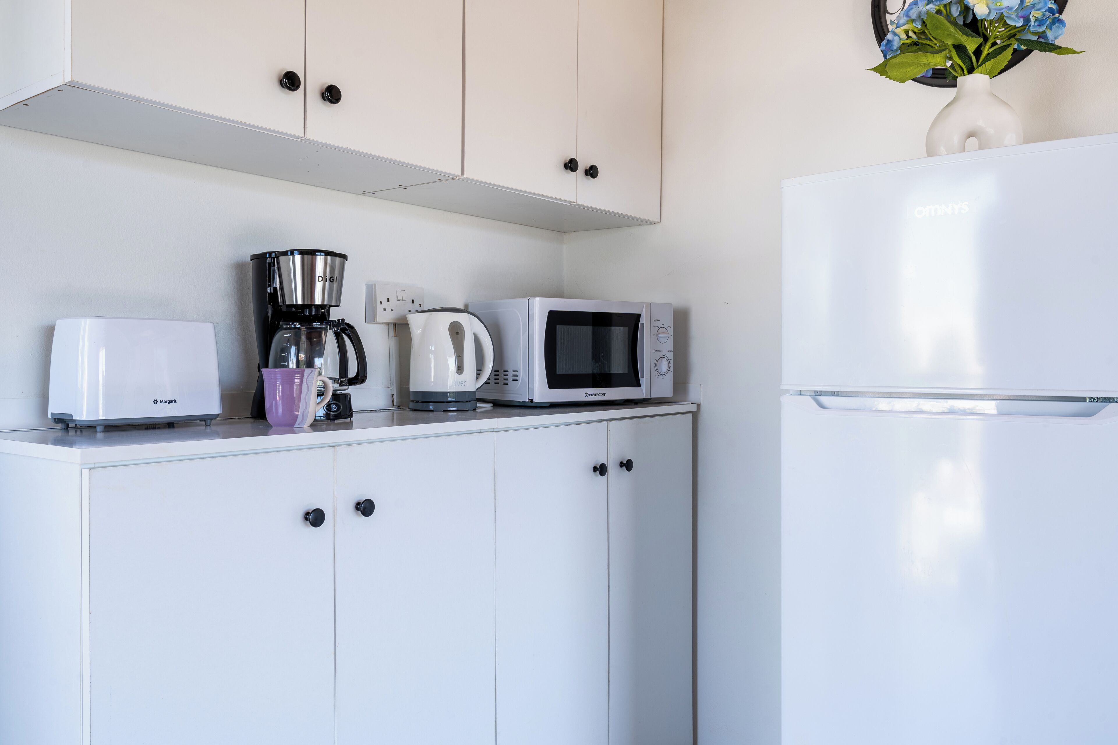 Una cucina con mobili bianchi, un frigorifero bianco, una macchina da caffè, un forno a microonde e un tostapane.