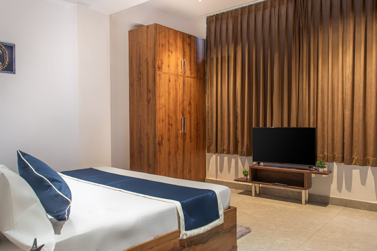 A bedroom with a bed, a wooden wardrobe, a television on a stand, and brown curtains.