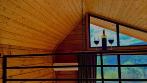 A cozy room with wooden slats on the ceiling, a large window with a view of the outdoors, a bed with a dark blanket, and a small table with a bottle and glasses. - Sioni Hillside Cottages (Sioni)