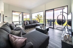 A modern living room with a sectional sofa, a coffee table, and a hanging chair. Large windows offer a view of the outdoors. - Tranquil Luxury Stay with Rice-Field & Ocean Views, Private Pool+Bar, Chef & BBQ (Bali)