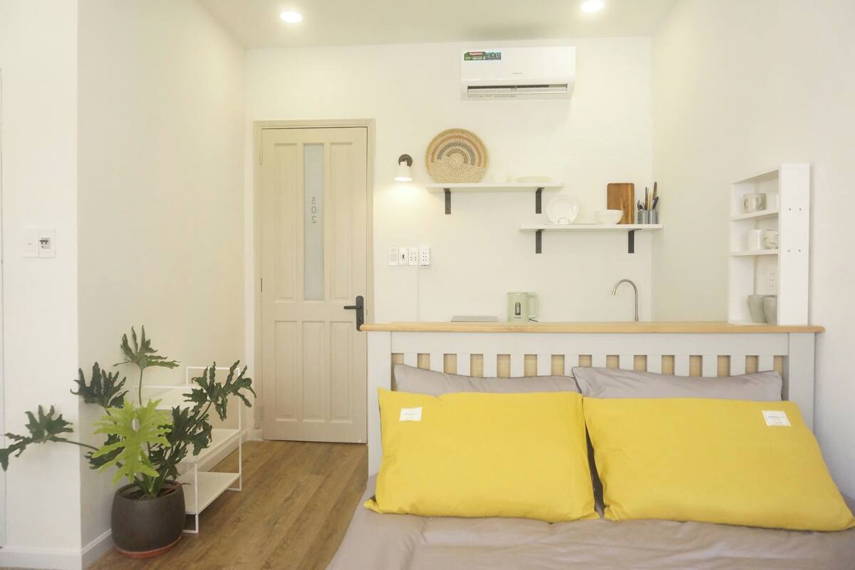 A compact room with a bed, a small kitchenette, a potted plant, and a door.