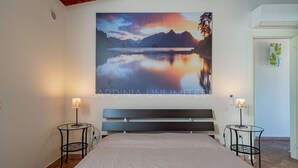 A bedroom with a large bed, two bedside tables, a bench, a painting of a mountain landscape, and a door with a green leaf pattern. - Villa Tramas de Luna - Sardinia Unlimited (Capo Coda Cavallo)