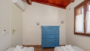 A bedroom with two beds, a blue dresser, a window with curtains, and a thermostat on the wall. - Villa Tramas de Luna - Sardinia Unlimited (Capo Coda Cavallo)
