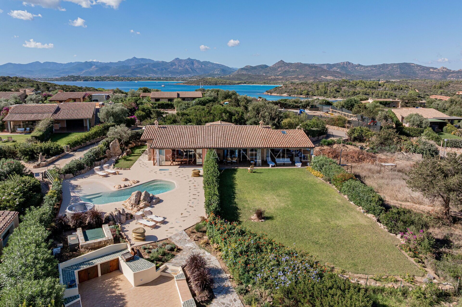 A large villa with a swimming pool, surrounded by lush gardens and a view of the sea and mountains.