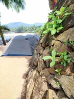 Una carpa montada en una playa arenosa con un muro rocoso y una palmera al fondo.