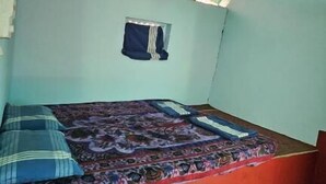 Ein kleines Zimmer mit einem Einzelbett, einem grünen Wellblechdach, einem Deckenventilator und einem kleinen Fenster mit Vorhängen.