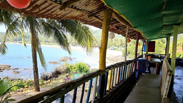 Blick auf einen Strand von einer überdachten Fläche mit Holzwand, einer Palme und Meer.
