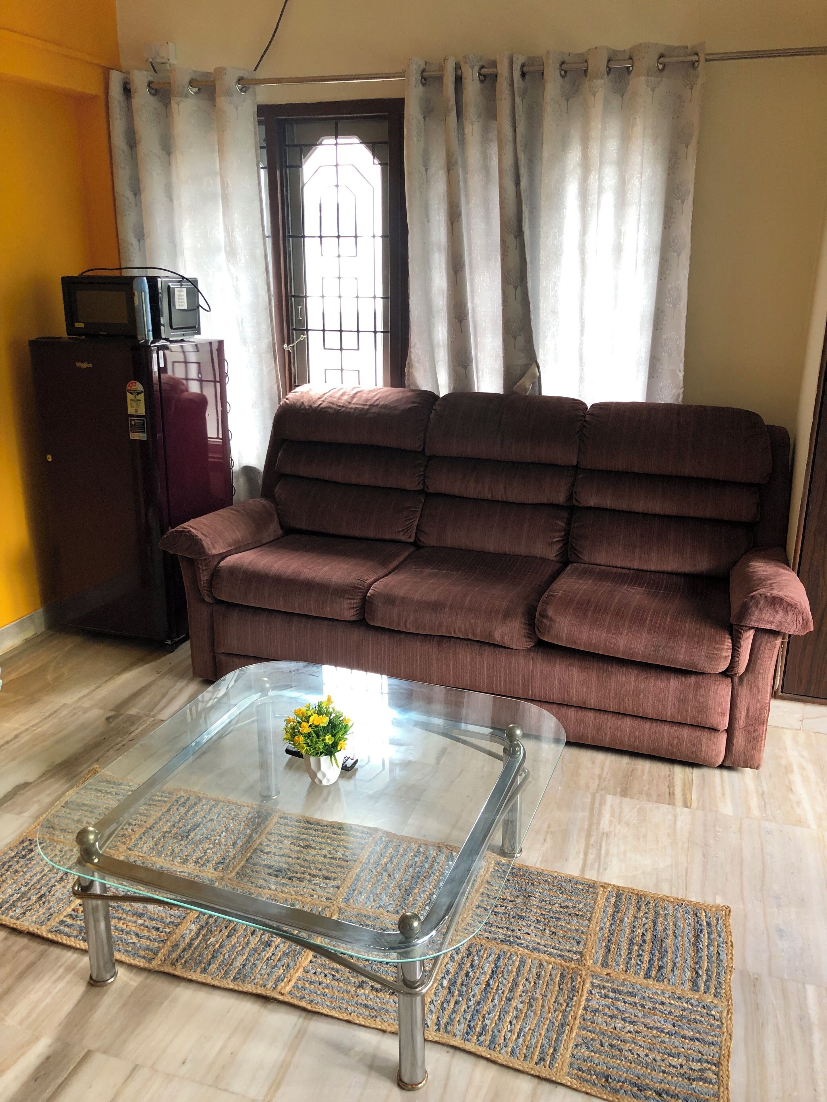 A living room with a brown sofa, a glass coffee table with a vase of flowers, a small refrigerator, and a door with a window.