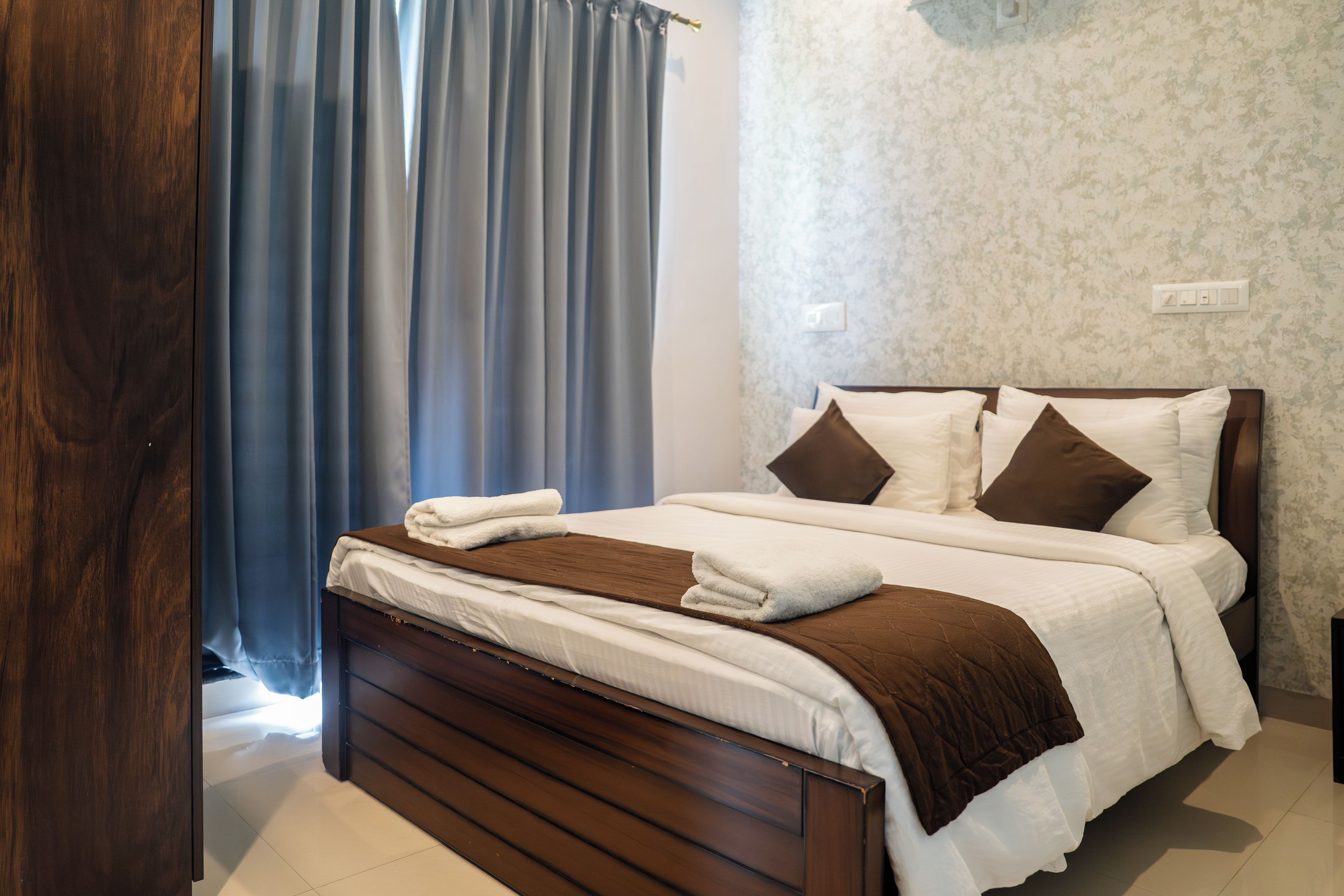 A neatly made bed with white and brown bedding, a wooden headboard, and a window with blue curtains.