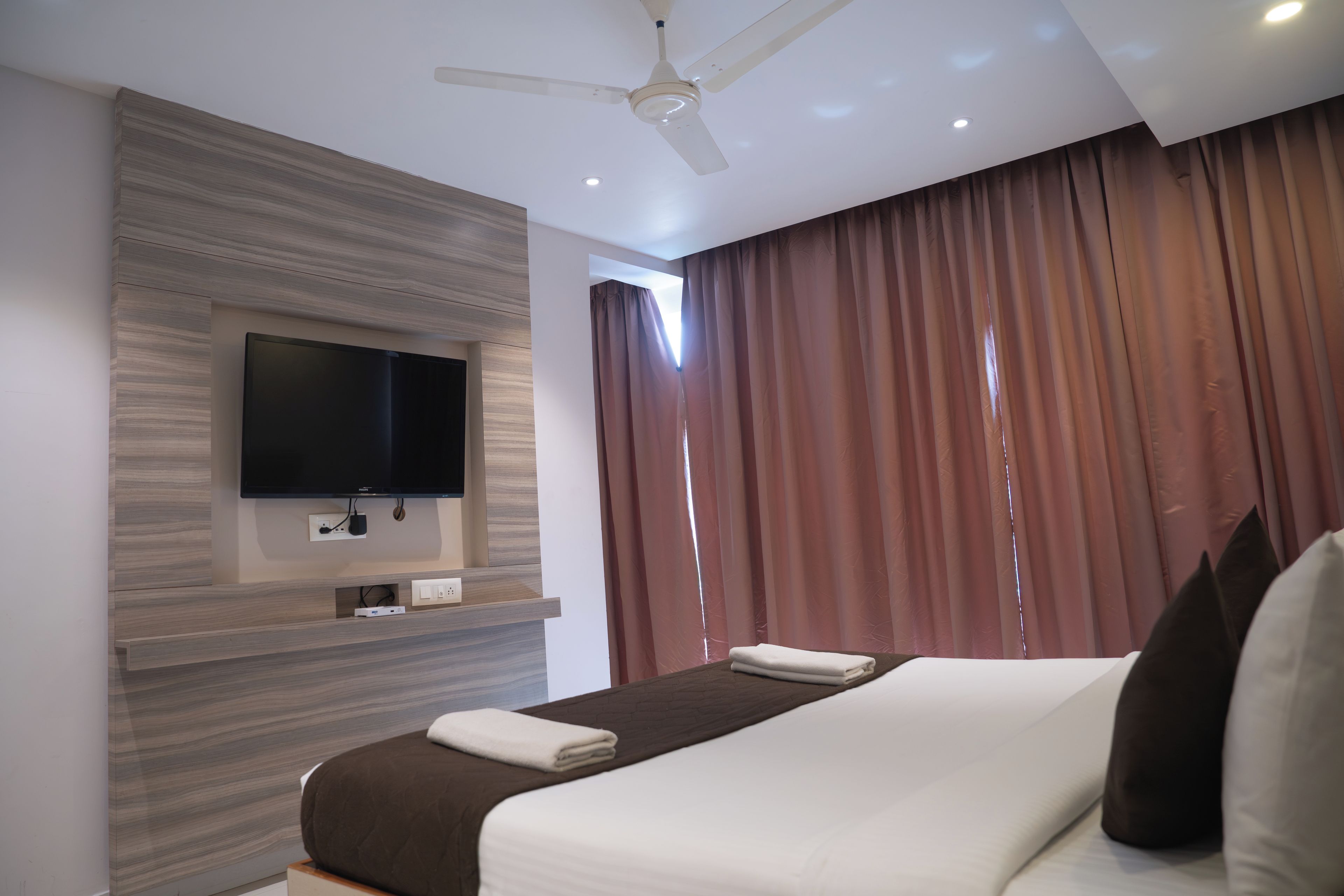 Habitación de hotel con televisión de pantalla plana montada en una pared de paneles de madera, cama con cabecera marrón y ventilador de techo.