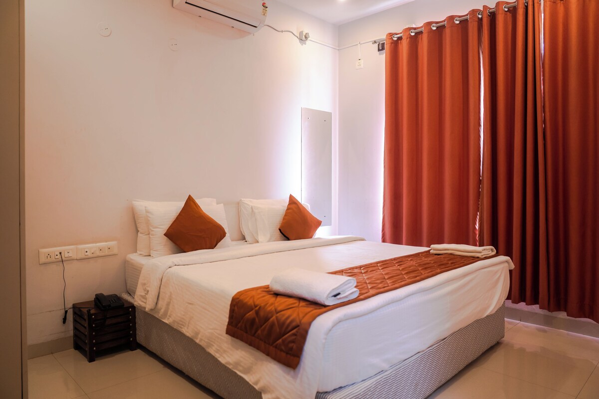 A hotel room with a bed, a bedside table, an air conditioning unit, and a window with curtains.