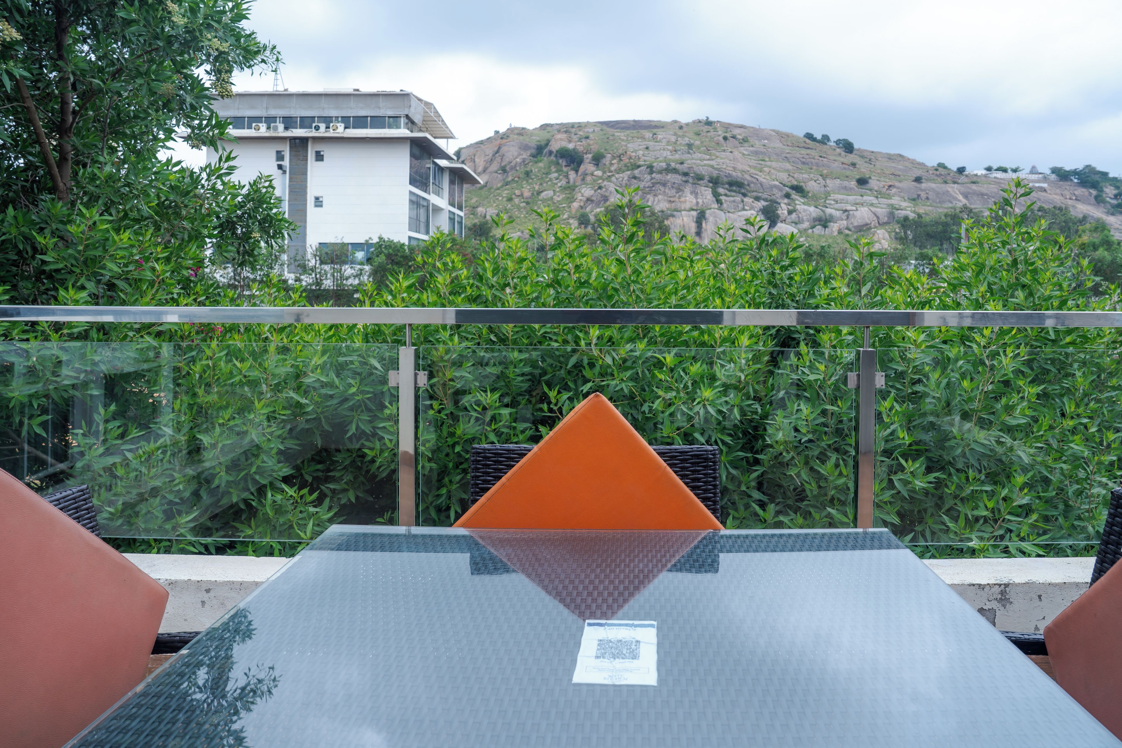 A modern outdoor seating area with a glass table and metal chairs, overlooking a mountainous landscape and a building.