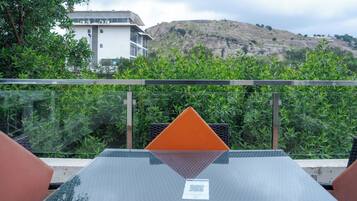 A modern outdoor seating area with a glass table and metal chairs, overlooking a mountainous landscape and a building.
