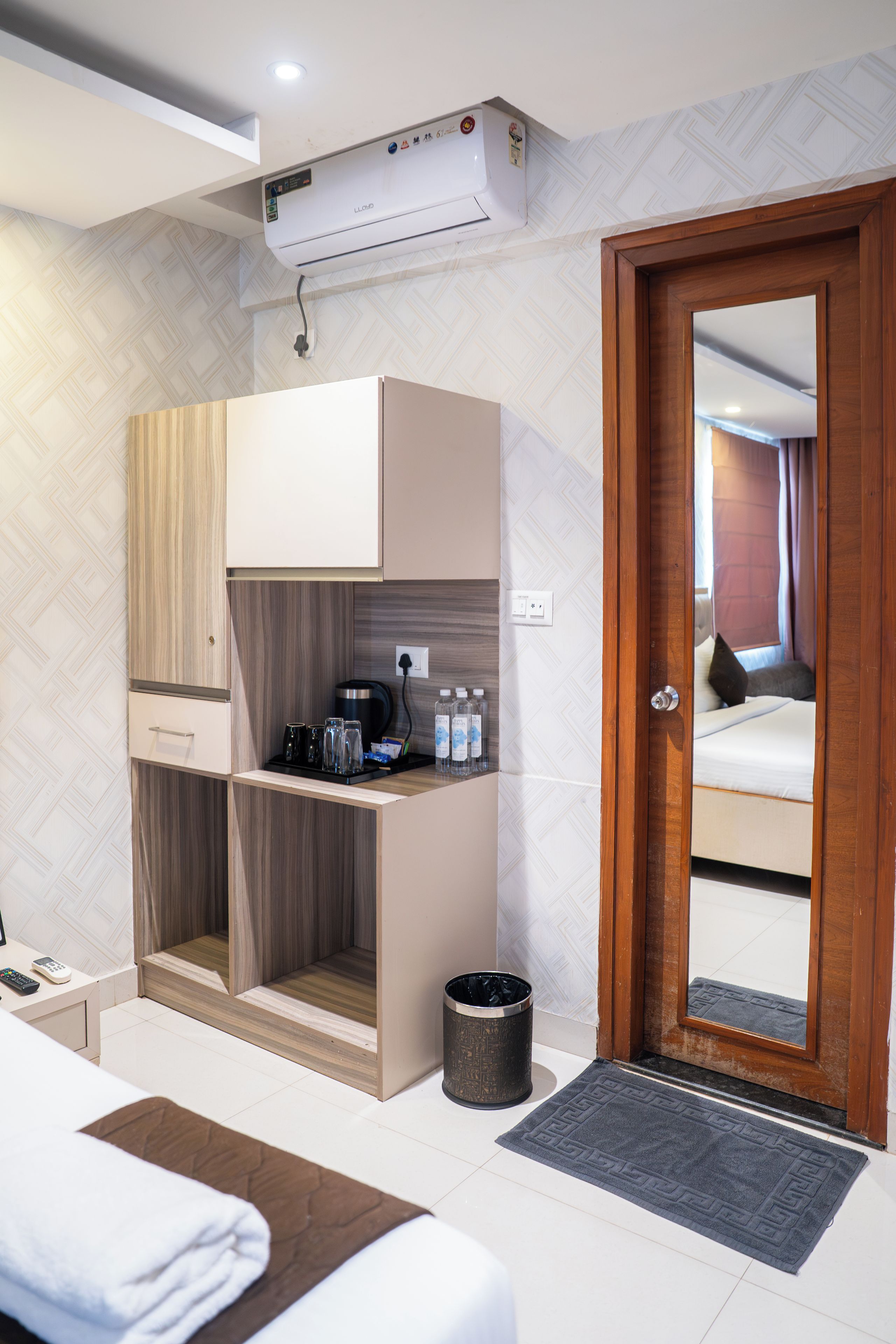A compact hotel room with a kitchenette, a bed, a bedside table, a trash bin, and a door leading to a bathroom.