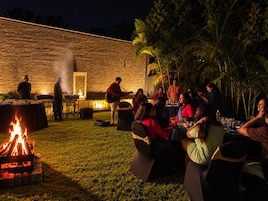 Reunión al aire libre con un brasero, mesas y personas cenando.