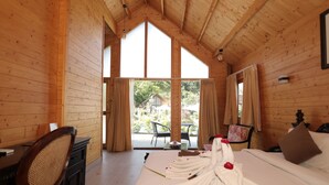 Una habitación de madera con una cama, un escritorio y una silla. Hay una ventana con cortinas y vista a follaje exterior.
