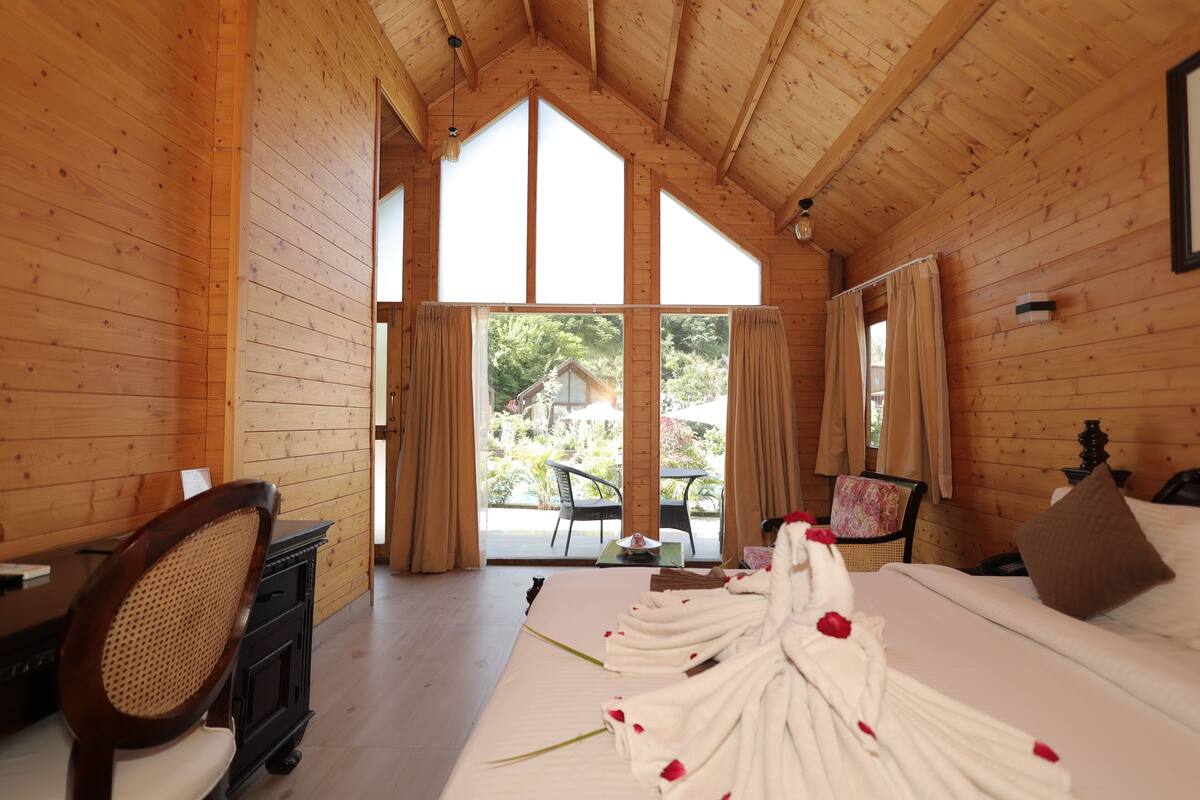 A wooden room with a bed, a desk, and a chair. There is a window with curtains and a view of greenery outside.