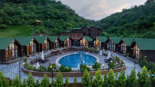 Un hotel con piscina central, varias habitaciones con techos verdes y un telón de fondo montañoso.