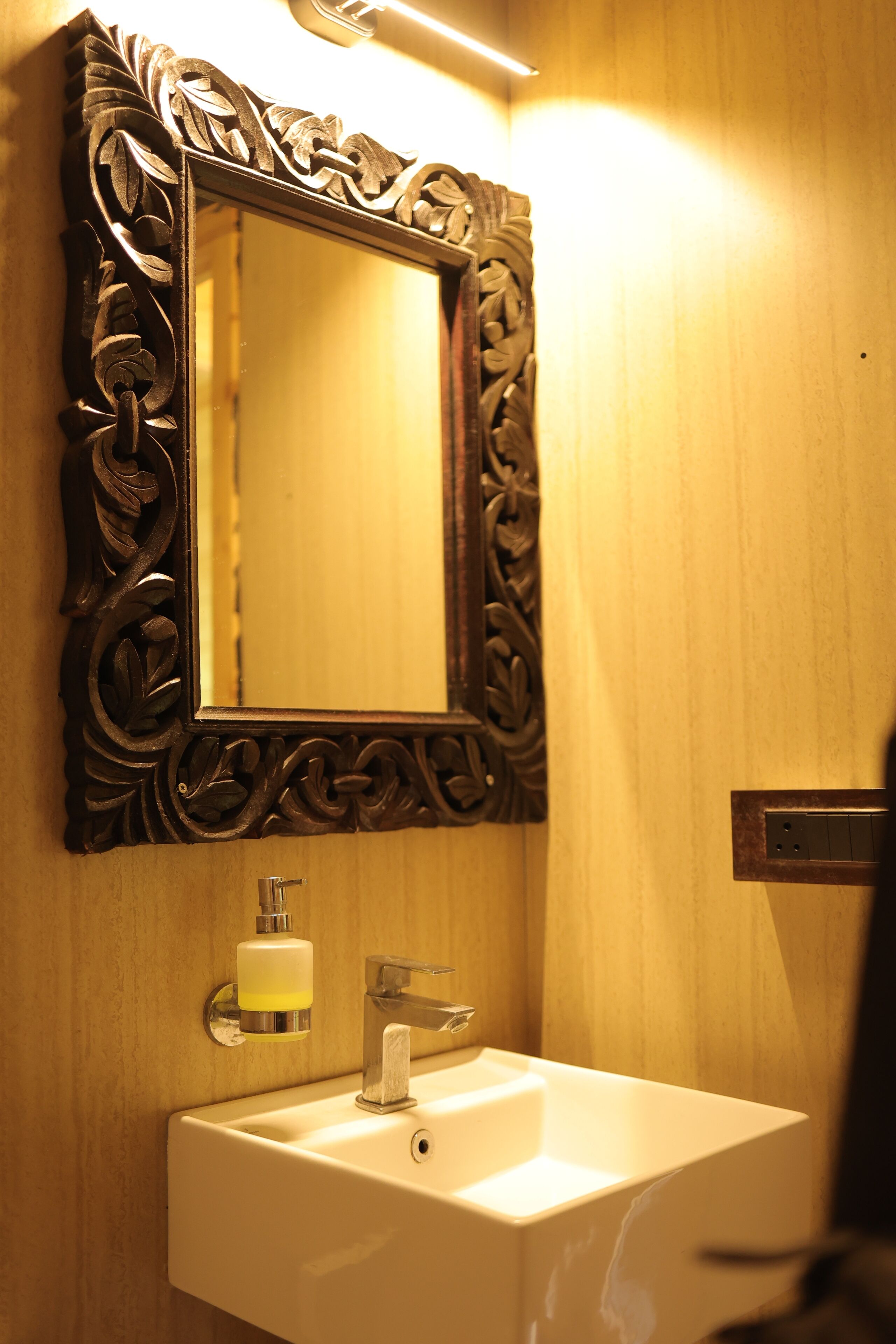 Un baño con pared de madera, un lavamanos blanco, un espejo con marco ornamentado y un dispensador de jabón.