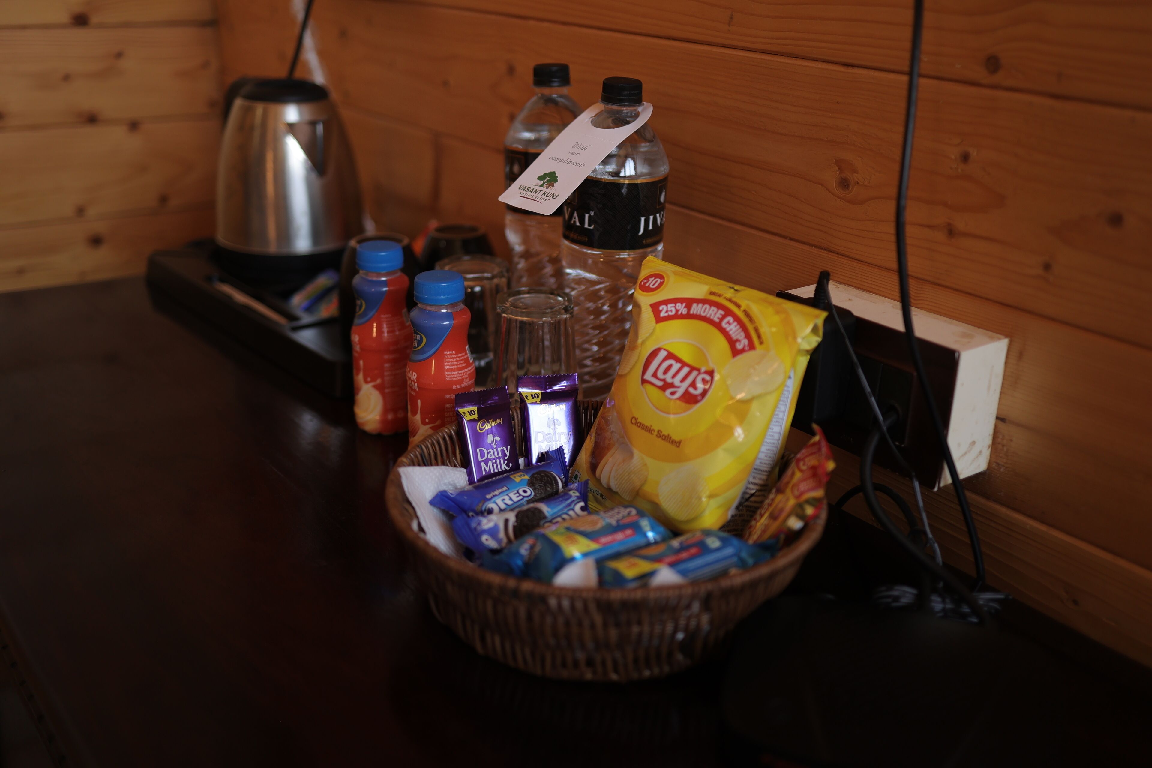 A wooden table with a basket containing snacks, a bottle of water, a bottle of juice, a bottle of milk, and a kettle.