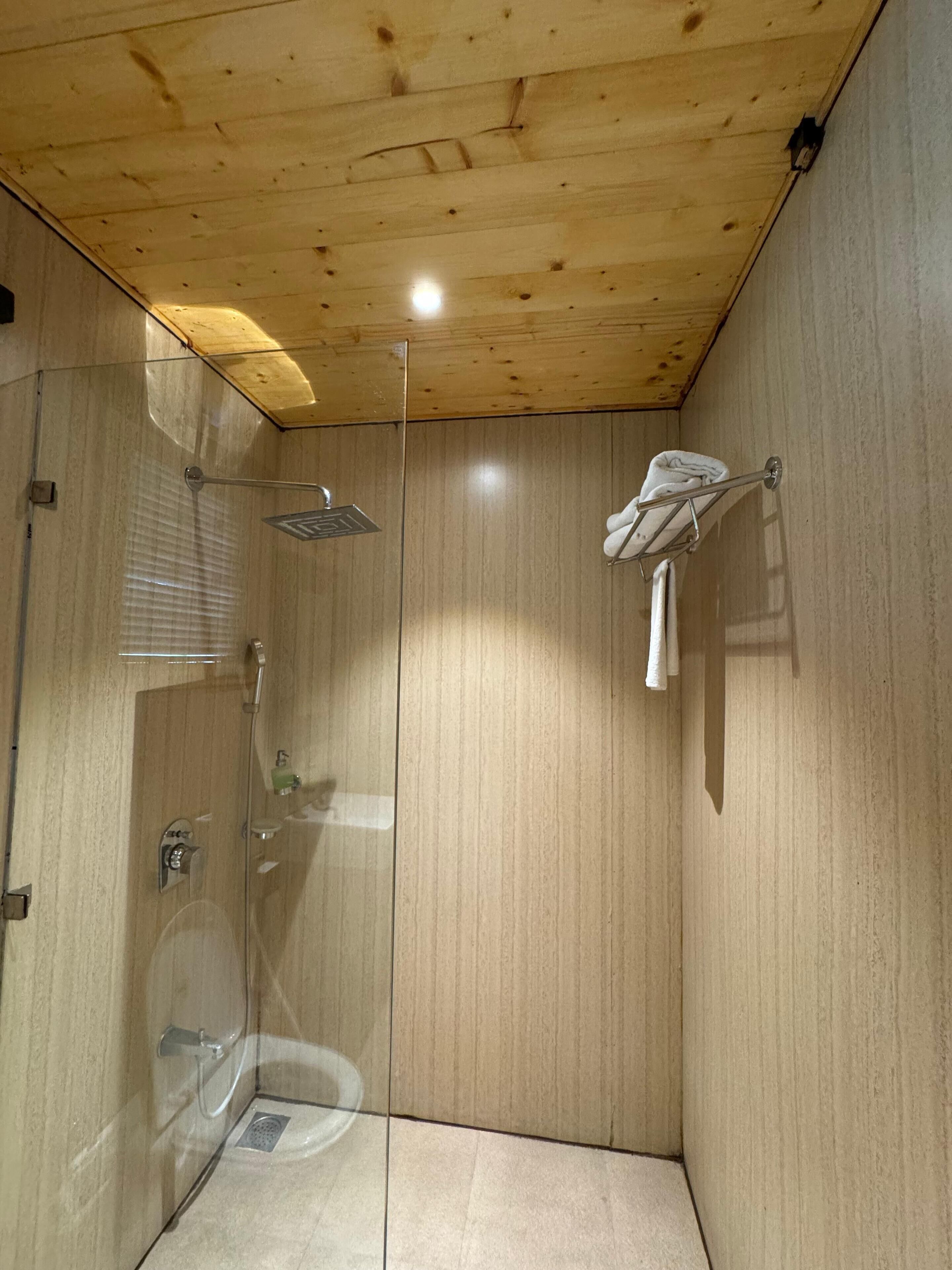 Un petit espace douche fermé, avec un plafond et des murs en bois, une porte en verre et un porte-serviettes sur lequel est accrochée une serviette.