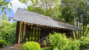 Ein hölzernes Pavillon mit geneigtem Dach, umgeben von üppiger Vegetation und einer gepflegten Hecke.