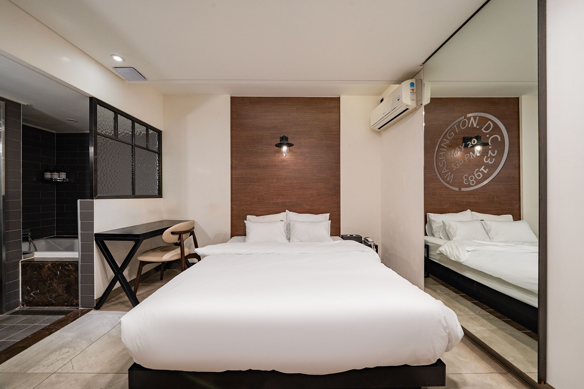 A hotel room with a large bed, a desk with a chair, a small bathroom, and a wall clock.