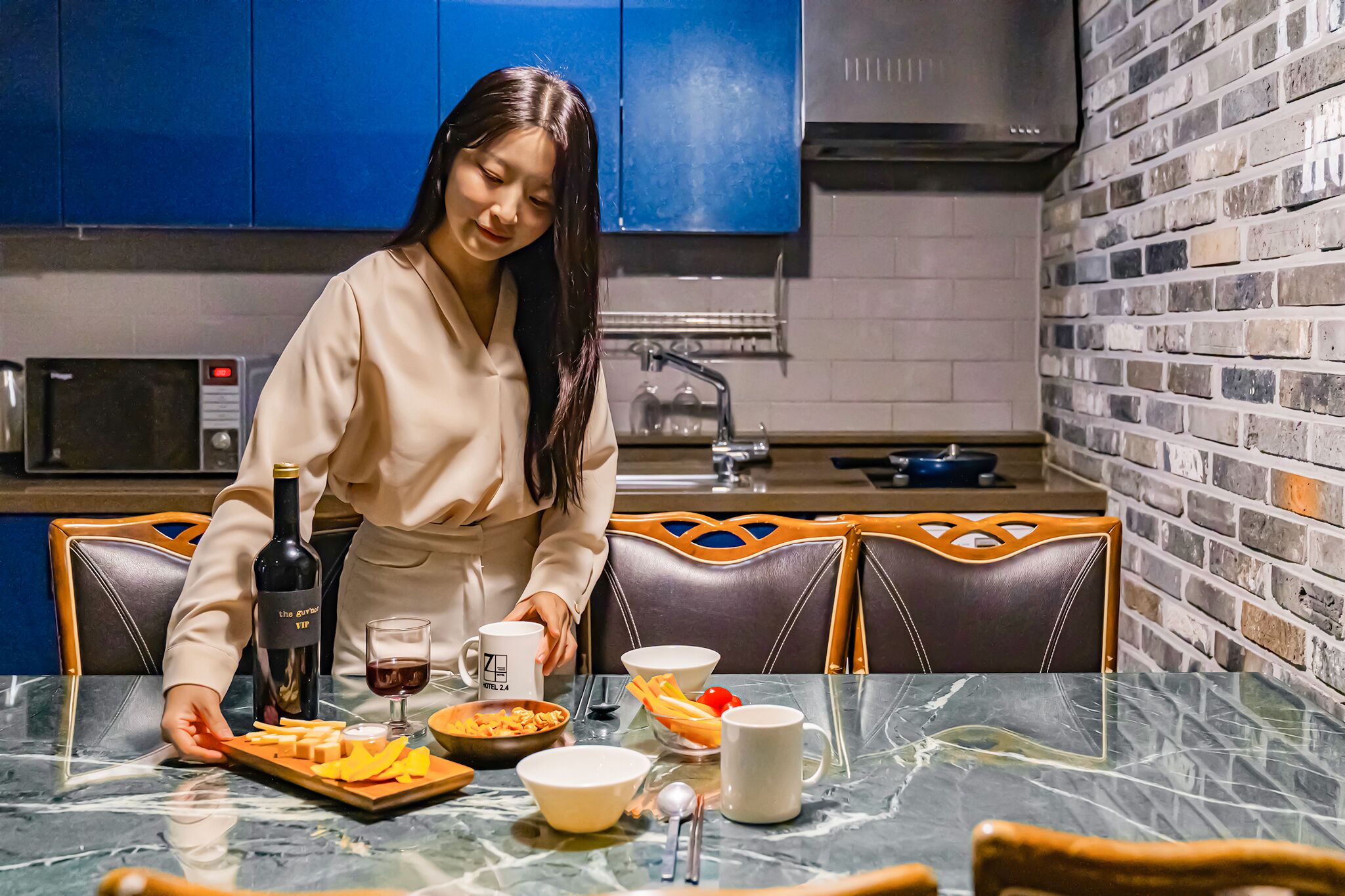 A person in a beige outfit arranging a meal on a glass table with a bottle of wine, bowls, and utensils.