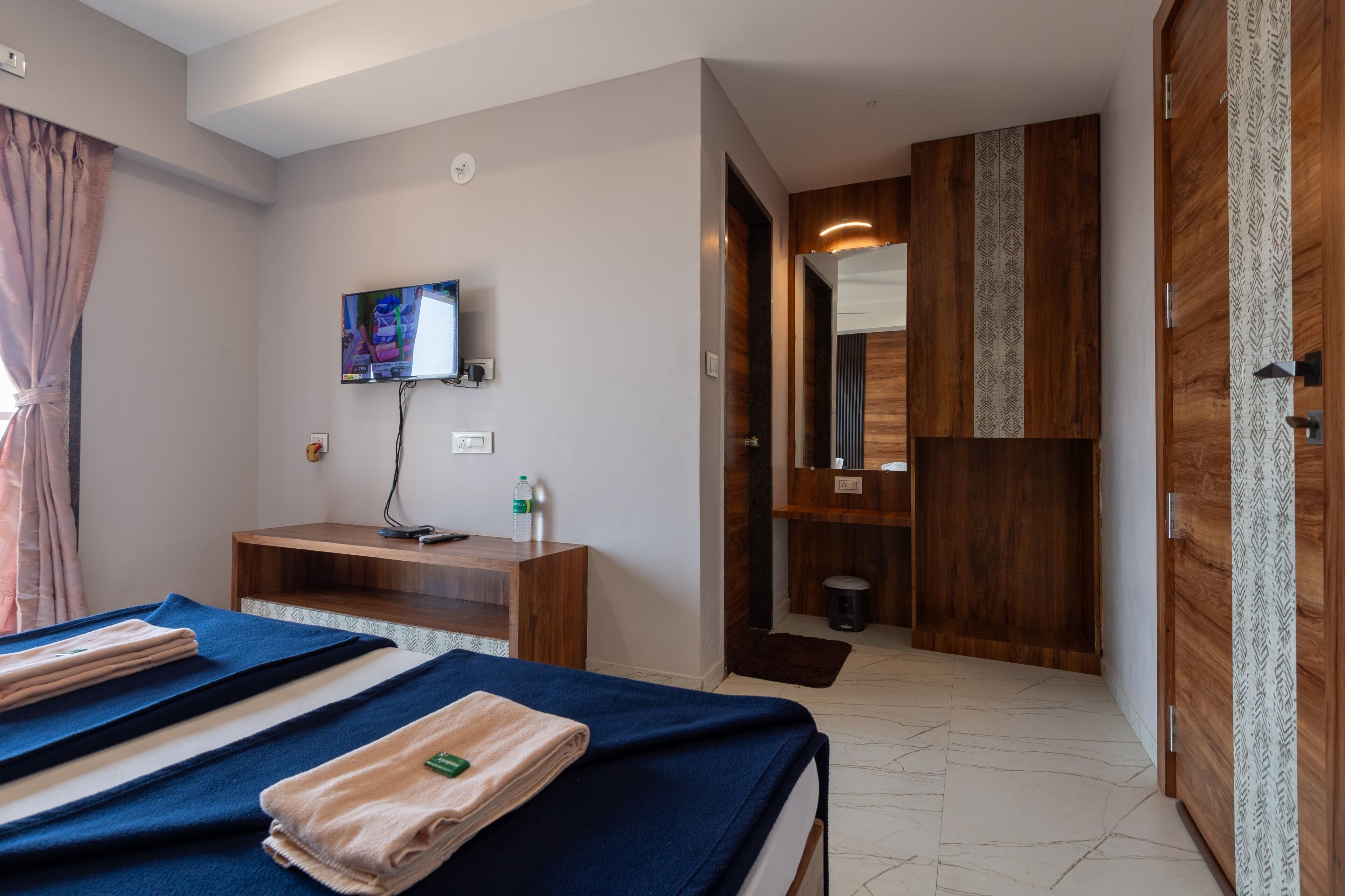 A hotel room with a bed, a TV mounted on the wall, a wooden desk, and a mirror.