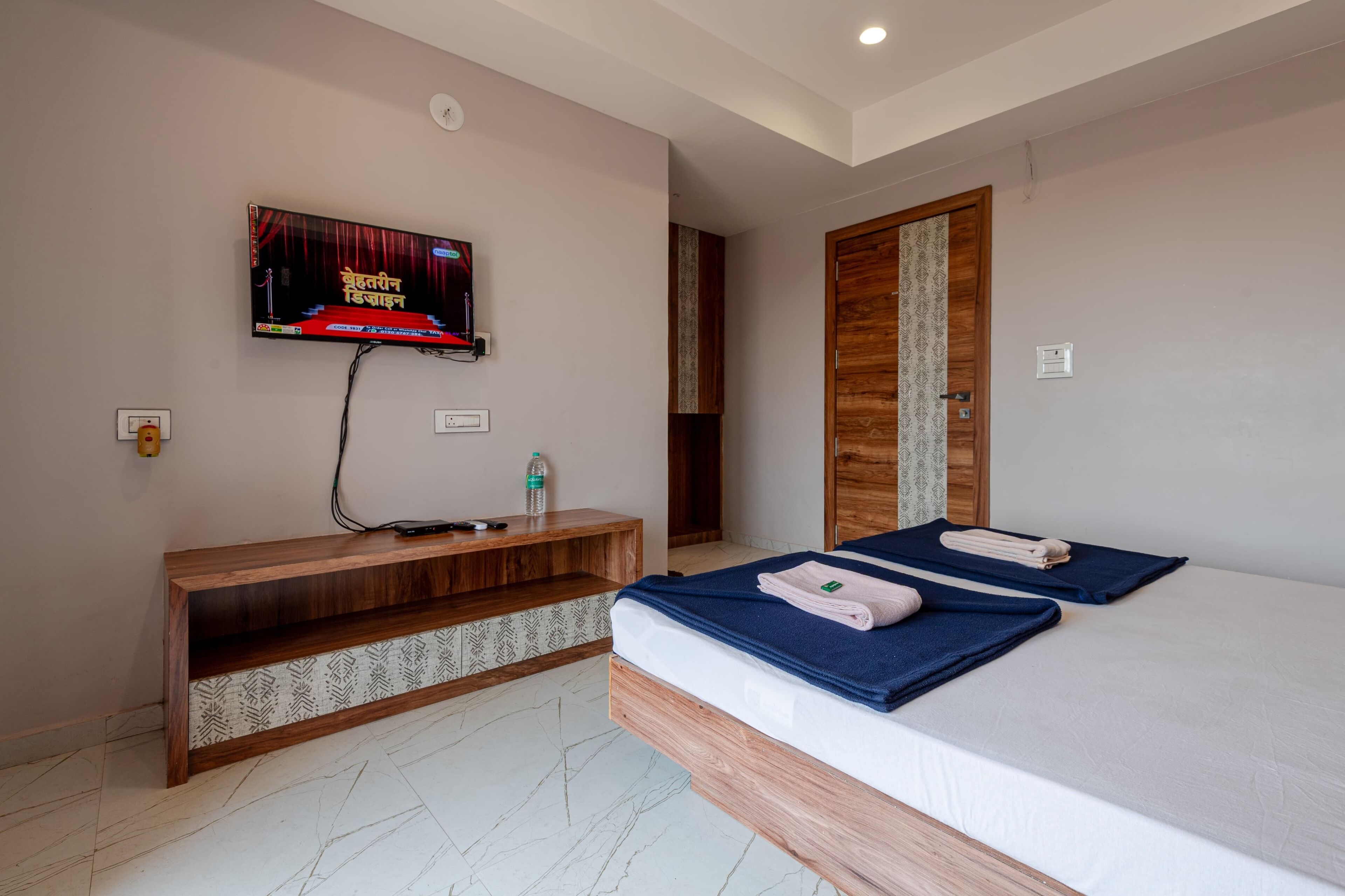 A hotel room with a bed, a TV mounted on the wall, a wooden console table, and a door with a patterned curtain.