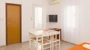 Ein kleiner, hell erleuchteter Raum mit einer Holztür, einem weißen Tisch mit Stühlen, einem an der Wand befestigten Fernseher und einem Fenster mit durchsichtigen Vorhängen.