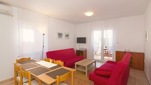 Een woonkamer met een rode bank, een houten eettafel met stoelen, een televisie en een raam met gordijnen.
