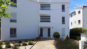 Un immeuble résidentiel moderne à plusieurs étages, doté d’une entrée centrale, d’un petit espace jardin et d’une allée pavée.