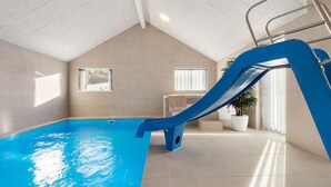 Una piscina interior con un tobogán azul, paredes de azulejos y techo inclinado.