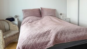Un lit bien fait, recouvert d’une courtepointe rose matelassée et agrémenté de deux oreillers, une chaise en osier, une table de chevet avec une lampe et une petite table de nuit sur laquelle repose un globe terrestre.
